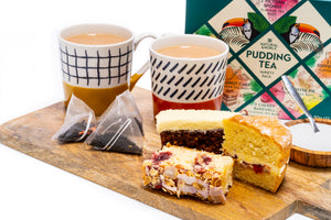 Two cups of pudding tea with a selection of pudding tea bags, slices of cake, and a box of pudding teas on a wooden serving board.
