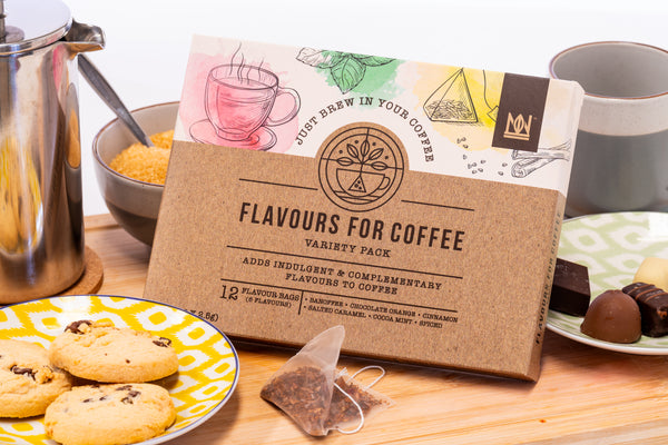 Box of 'Flavours for Coffee' variety pack on a wooden table with cookies and tea bags.