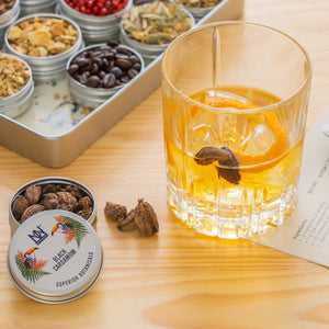 Glass of whisky infusing with cardamom and orange peel, adjacent to a selection of dried ingredients.