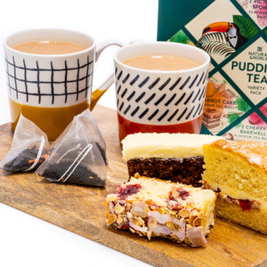 Two cups of pudding tea with a selection of pudding tea bags, slices of cake, and a box of pudding teas on a wooden serving board.