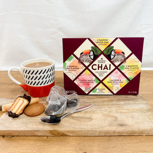 A variety pack of Chai tea bags displayed alongside a cup of tea and cookies on a wooden surface. The packaging is colourful and includes six different Chai flavors.