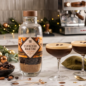 A bottle of a Natural & Noble DIY cocktail kit cocktail mix labelled 'Espresso Martini Original' stands on a marble countertop, with two prepared espresso martinis in coupe glasses, coffee beans, almonds, and a blurred espresso machine in the background.