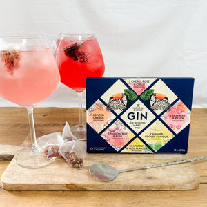A variety pack of gin infusion bags displayed alongside two glasses of gin tonic with garnishes, and a spoon on a wooden board. The packaging is colorful and includes text detailing the different flavor blends.