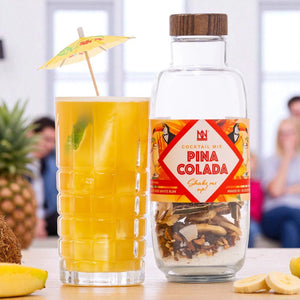 Pina Colada cocktail mix bottle with a glass of cocktail and pineapple, banana, and coconut on a blurred background.