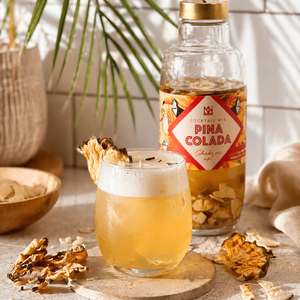Tropical Pina Colada Alongside Natural & Noble Cocktail Kit, Surrounded by Pineapple and Coconut Pieces