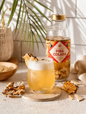 Tropical Pina Colada Alongside Natural & Noble Cocktail Kit, Surrounded by Pineapple and Coconut Pieces