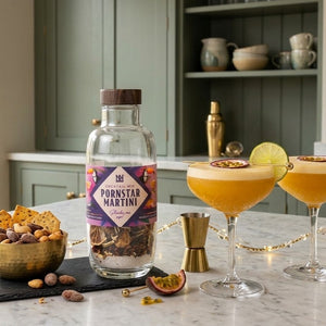 A bottle of a natural & noble DIY Pornstar Martini cocktail mix standing on a marble countertop in a kitchen setting, with two prepared cocktails in coupe glasses, nuts, and passion fruit nearby.
